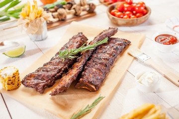 Spicy hot grilled spare ribs from a summer BBQ served with chips, corn and fresh tomatoes on an old wooden cutting board