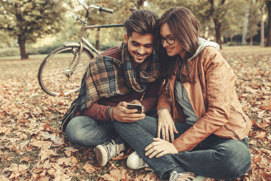 Young couple sitting outdoors at the park on beautiful autumn day.They sitting leaves and using smart phone.
