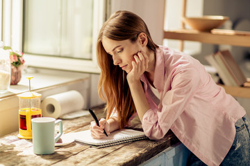 Portrait of young girl writing in notebook, standing in the kitchen. Female in casual clothes...