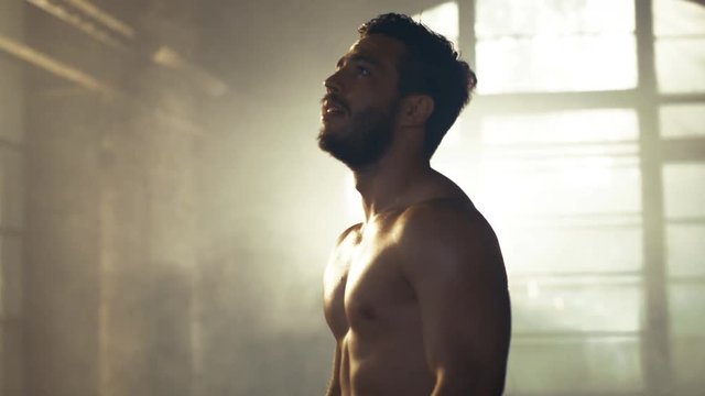 Handsome Shirtless Man with Naked Muscular Torso with Visible Six Pack Stands Resting after Bodybuilding Exercise, He Wipes Sweat from His Forehead. He's in the Middle of Abandoned Factory Building. 