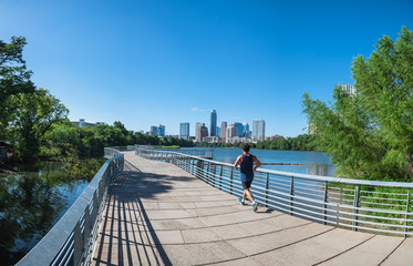 Panorama view Downtown Austin, Texas, US along Colorado River at daytime with cloud blue sky. View from Ann and Roy Butler Hike-and-Bike Trail and boardwalk at Lady Bird Lake, unidentified man running - obrazy, fototapety, plakaty