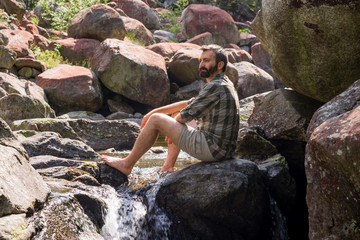 Young barefoot adult man seated on a rock on the top of little waterfall refresh himself on mountain river in the morning in summer outdoor.