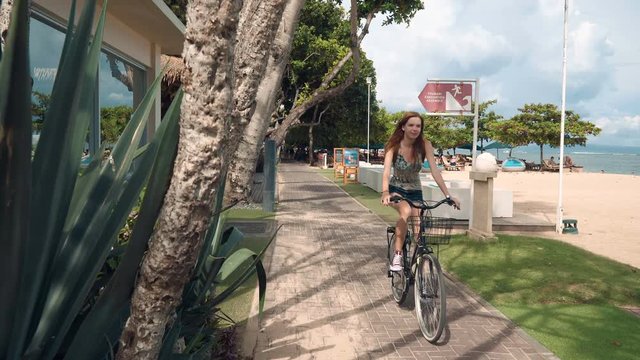 Young caucasian woman tourist in top and jeans shorts rides bicycle on Sanur beach road. Shot with Sony a7s and Atomos Ninja Flame on sunny day