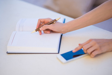 Photo of opened notebook in which woman notes something with help of pen and keeps a phone