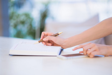 Photo of opened notebook on table in which woman notes something with help of pen and uses a phone
