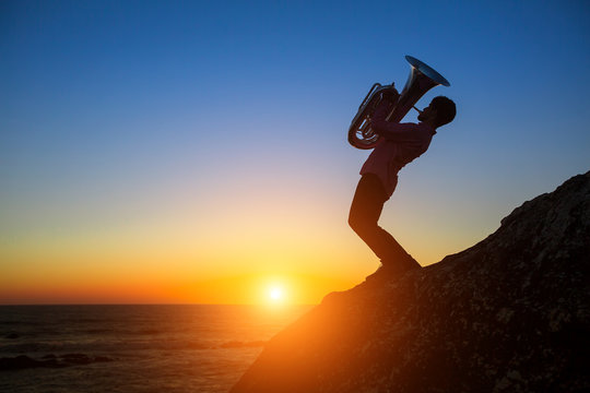 Silhouette of musician with Tuba on rocky sea coast during amazing sunset.