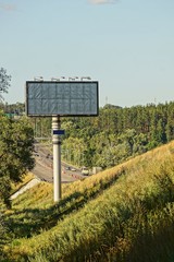 Большой рекламный щит на трубе в траве у дороги с машинами на фоне неба