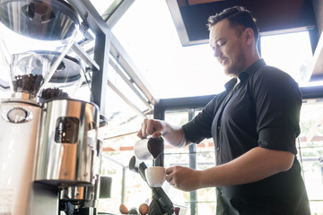 Side view of a young serious bartender pouring fresh milk into a cup of coffee behind the bar counter in a trendy cafeteria