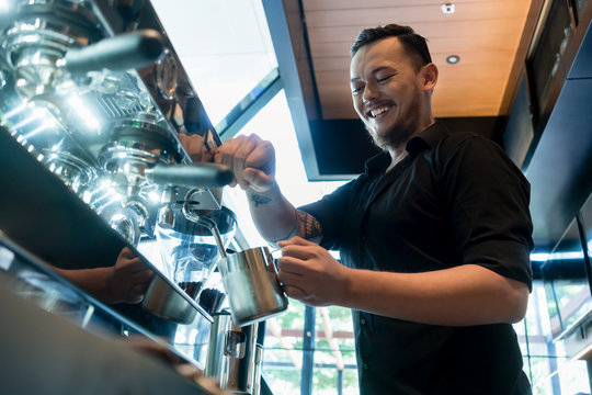 Low angle view of a young cheerful barista wearing black shirt while preparing coffee at an automatic machine in a modern coffee shop
