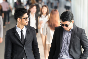 Young Asian Business men walking with his colleague outside the office and talking about their work . One Businessman looking at his mobile phone. Both of them wear sunglasses