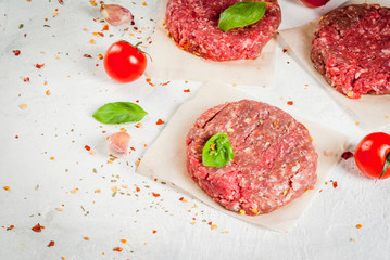 Fresh raw home-made minced beef steak burger with spices, tomatoes and basil, on a white stone concrete table, copy space
