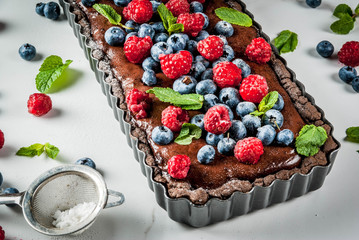 Summer homemade baked pastry. Chocolate cake tart with chocolate cream, fresh raw berries blueberry raspberry, decorated with mint leaves, powdered sugar. On white marble table, copy space