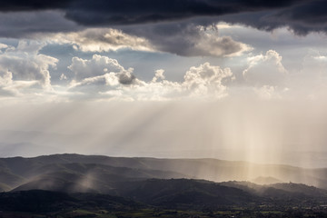 Spectacular view of sun rays and rain over some mountains and hills, benath a moody and cloudy sky