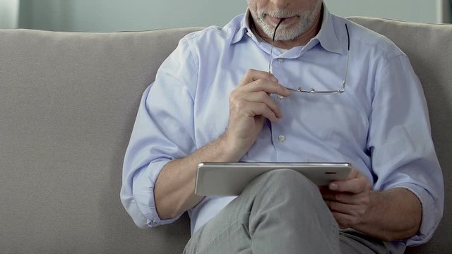 Elderly man holding tablet on lap, planning and booking retirement trip, closeup