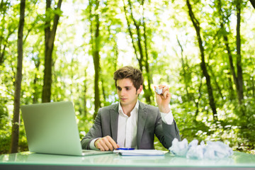 Young handsome business man at work table office with laptop in green forest with crumpled papers feeling exhausted angry and sad no ideas. Business concept