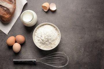 Bowl with white flour, eggs, bread, milk in jar and whisk on kitchen table