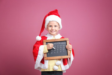 Cute little girl in Santa Claus suit with chalkboard counting days until Christmas, on color background