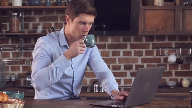 Young businessman have breakfast and working at home. Millennial man sitting in kitchen using laptop typing mail or surfing internet or reading news and drinking coffee in the morning.