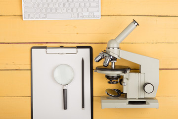Education and science concept - microscope, magnifying glass, blank clipboard, computer keyboard on the yellow desk