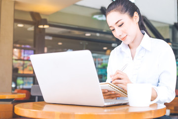 Business woman working in a computer and writing with a pen at a notepad in the office.