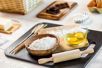 Baking ingredients in rural kitchen - dough recipe ingredients (eggs, flour, milk, butter, sugar) and rolling pin on wooden white table.