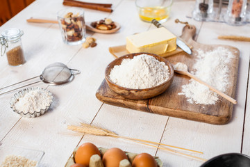 Baking ingredients in rural kitchen - dough recipe ingredients (eggs, flour, milk, butter, sugar) and rolling pin on wooden white table.