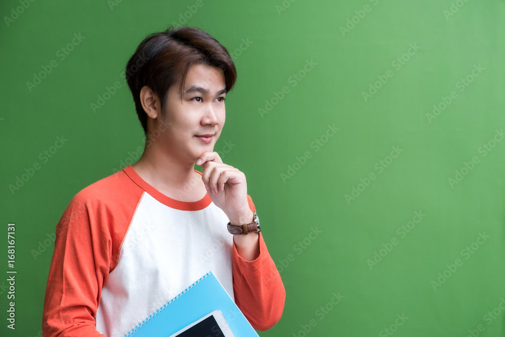 Poster young teenager man holding tablet computer and notebook and thinking action at green wall in univers