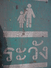 Old peeled white painted parent and child icon sign with Thai language word "Be careful" on green paved road background
