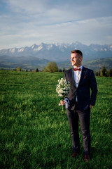 Groom with wedding bouquet stands on the field before the mountains