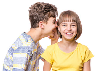 Fototapeta premium Friendship - teen boy whispering something to beautiful girl. Portrait of happy brother and sister, isolated on white background. Funny couple children - first love.