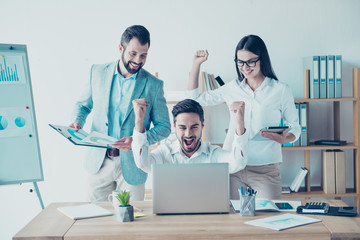 Yes! We did it! Success and team work concept. Team of three business partners with raised up hands with fists in light modern work station, celebrating the growth of their company