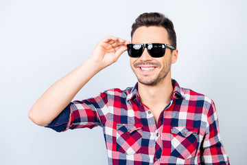 So cool and cheerful! Happy young brunet guy with stubble on vacation in a stylish sun protective spectacles,  wearing casual checkered shirt, holding his glasses