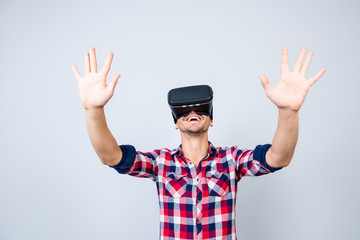 Excited young man is getting experience using VR-headset glasses of virtual reality gesticulating with his hands, in casual chekered wear, isolated on light background