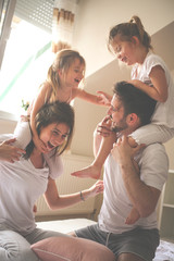 Family spending free time at home. Daughters sitting on the shoulders of their parents. Fun on bed....