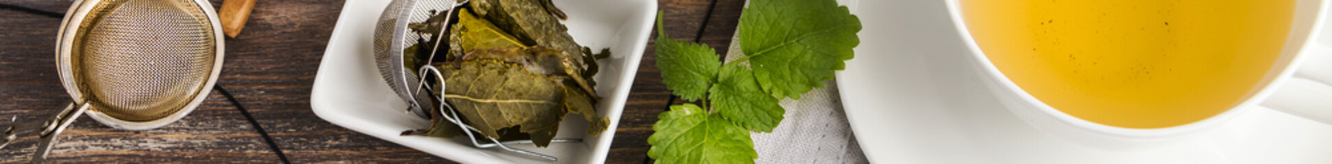Cup of green tea, honey, tea leaves, green mint leaves, tea strainers of rustic wooden table. Top...