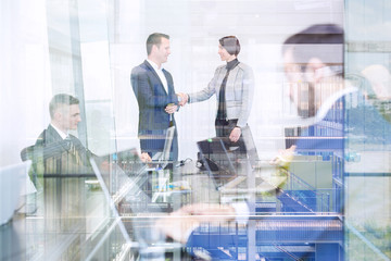 Sealing a deal. Business people shaking hands, finishing up meeting in corporate office. Businessmen working on laptop seen in glass reflection. Business and entrepreneurship concept.