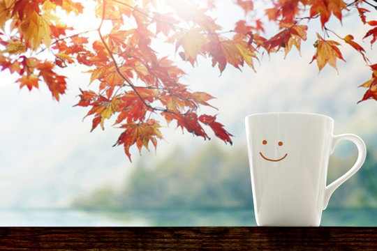 Happiness and Relaxation moment in Fall Season Concept, Happy smiley face Coffee Cup with Autumn Foliage Maple Leaf, Blurred Lake and Mountain  as background