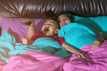 Fototapeta na wymiar Mother and son sleeps on the bed and holds the TV remote and smiles happily, concepts of technology and entertainment.