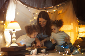 Reading and family games in children's tent. Mother and two twins daughters with books and flashlight before going to bed.