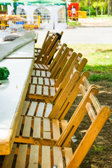 Fototapeta na wymiar Long tables in garden row of wooden chairs. Preparation for outdoor summer picnic or public fest. Green grass, sunlight flecks, leisure. Lifestyle image.