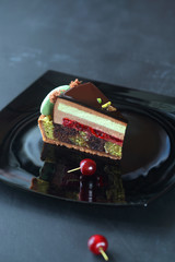 Piece of Contemporary Chocolate, Cherry and Pistachio Layered Mousse Cake, on black plate, on dark background.