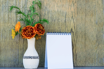 Abstract background, blank white sheet of paper in the notebook and a bright ceramic vase with a bouquet of orange flowers on background wooden wall vintage. Mock up