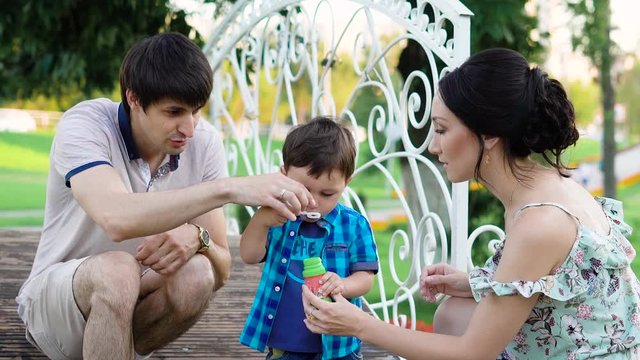 Young parents are teaching their son to blow bubbles, mum is holding a jar of liquid, dad is afraid to blow, baby likes a new game
