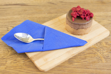Piece of cake with chocolate glaze and raspberries on a plate on a wooden background. Blue napkin with a spoon beside the cake.