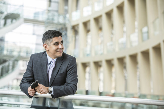 Mixed Race businessman leaning on railing in lobby holding cell phone