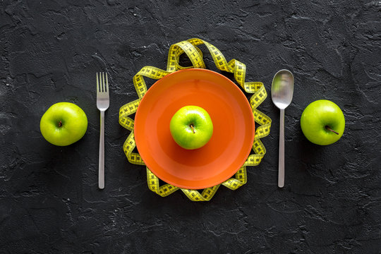 Slimming diet. Apple at plate and measuring tape on black background top view