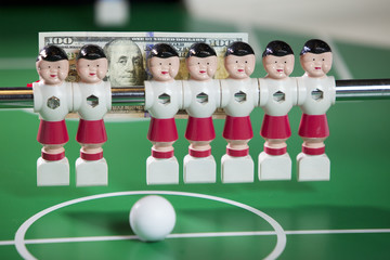 Sports and money concept-toy football players stand in the field and the banknote in hundred dollars on a background