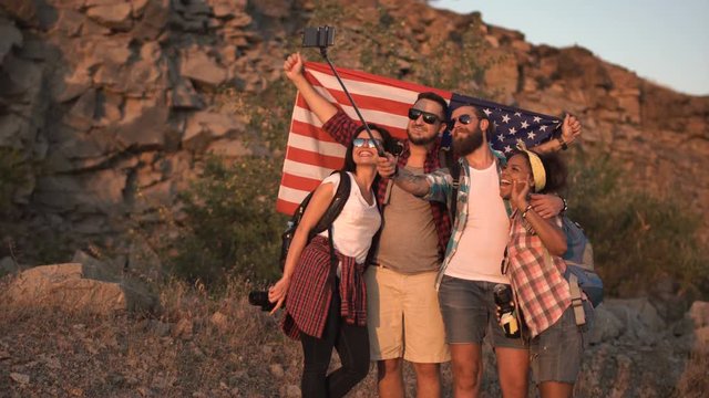 Multiracial group of friends posing with backpacks and American flag taking selfie on nature.