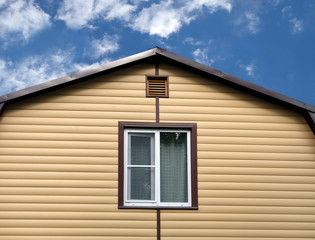 Top section of modern private country house wall covered with yellow siding and brown metal roof on blue sky with clouds on sunny day front view closeup