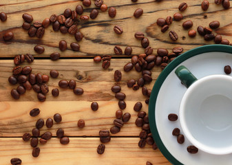 Roasted coffee beans scattered on the wooden table next to an empty cup on a saucer with a green rim in the right bottom corner